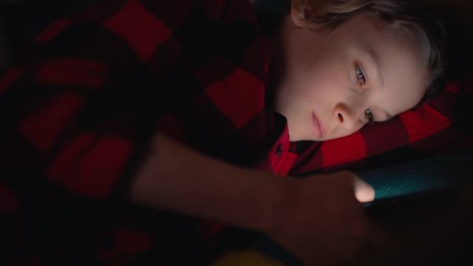 可爱的小男孩躺在黑暗房间的床上，在家使用智能手机。快乐的青少年在线浏览内容，在社交媒体上与朋友聊天，