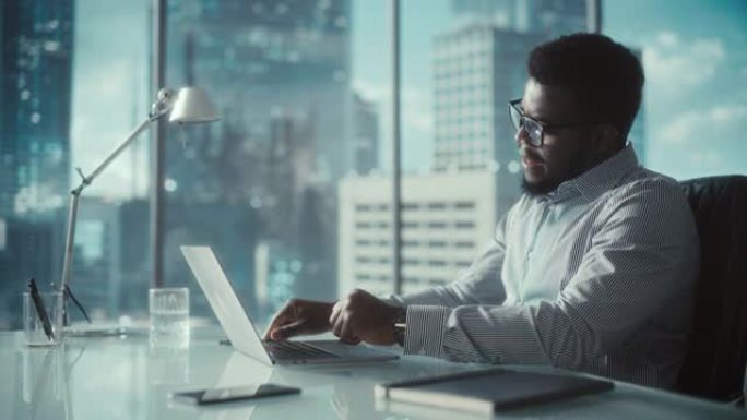 穿着条纹衬衫的自信商人坐在现代办公室的办公桌前，在窗户旁边使用笔记本电脑，可以看到摩天大楼。成功的财