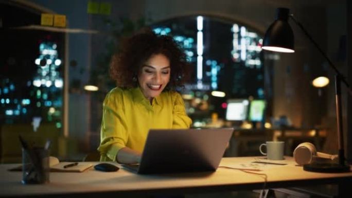 富有表现力的中东女性晚上坐在笔记本电脑上工作。项目经理收到有关她的业务提交的积极消息。兴奋的专家很高