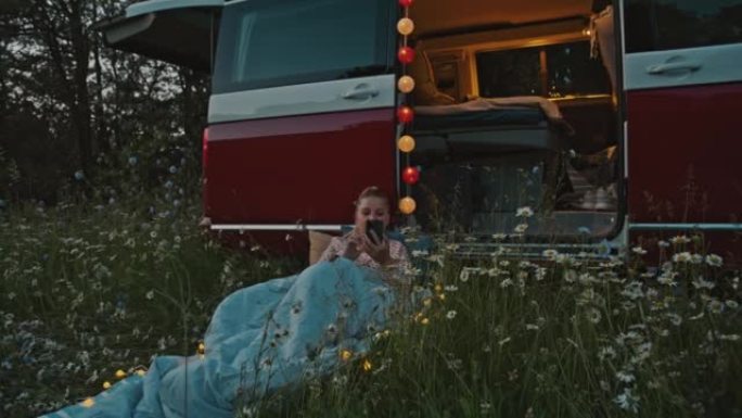 DS女人在黄昏时躺在停在草地上的露营者旁边时使用智能手机
