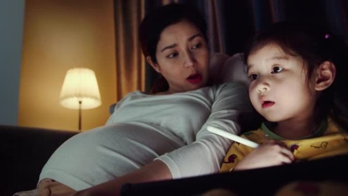特写程序员怀孕的母亲在笔记本电脑上编码，而女儿在晚上在家使用数字平板电脑。