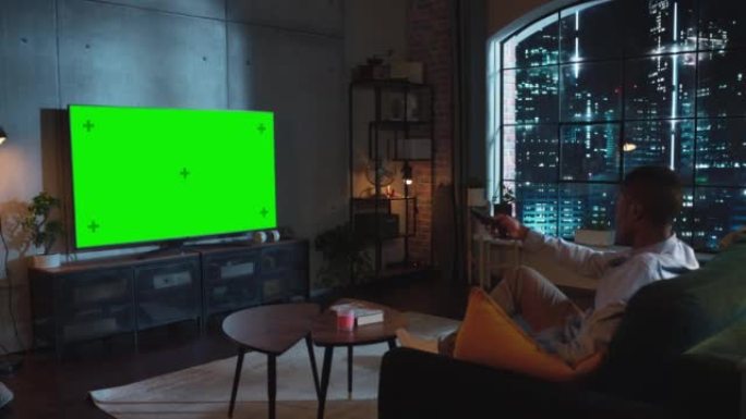 在阁楼客厅，非裔美国人坐在沙发上，打开带有控制器的绿屏模拟显示的电视。男性喜欢在家休闲。夜间射击
