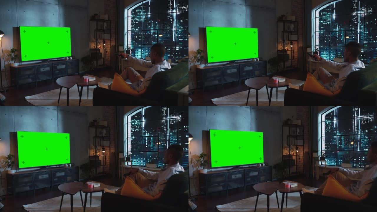 在阁楼客厅，非裔美国人坐在沙发上，打开带有控制器的绿屏模拟显示的电视。男性喜欢在家休闲。夜间射击