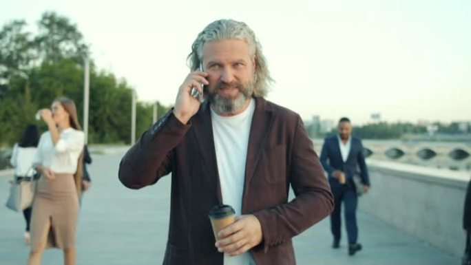 英俊成熟的商人沿着堤岸行走，拿着咖啡在手机上聊天