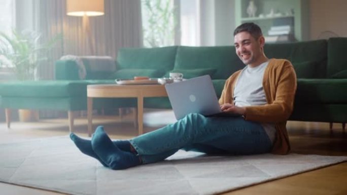 英俊的成年男子使用笔记本电脑，同时坐在舒适时尚的公寓的客厅地板上。有魅力的男人正在与工作中的朋友或同