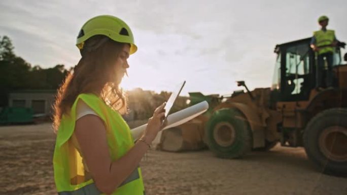 慢动作，手持一位黑发女采矿工程师在大型黄色推土机前使用平板电脑拍摄，男工程师站在上面