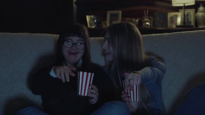 唐氏综合症的姐姐和少女在家里一起看电视时吃爆米花的电影镜头。概念: 技术，连接，残障，残障，娱乐，智