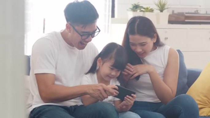 快乐的亚洲家庭爸爸，妈妈和女儿坐在家里房间的沙发上玩智能手机上有趣的在线游戏。