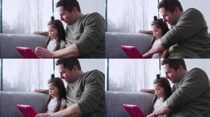 父亲和女儿坐在家里的沙发上一起在粉红色盒子里的数字平板电脑上玩耍