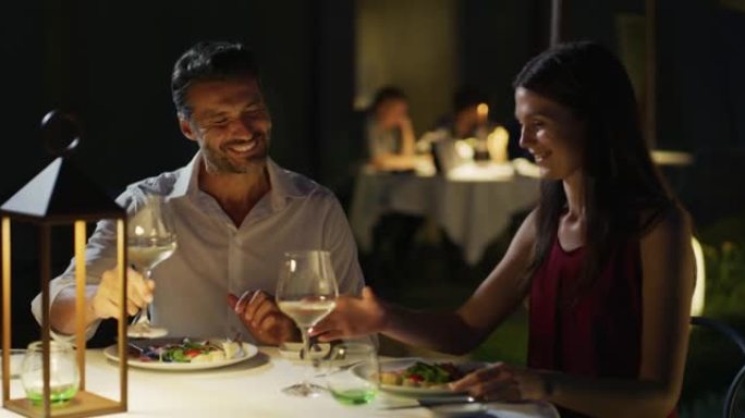 穿着优雅的幸福夫妇一起享受浪漫的晚餐，并在豪华餐厅的餐桌上用白酒杯欢呼，庆祝他们的周年纪念日和永恒的
