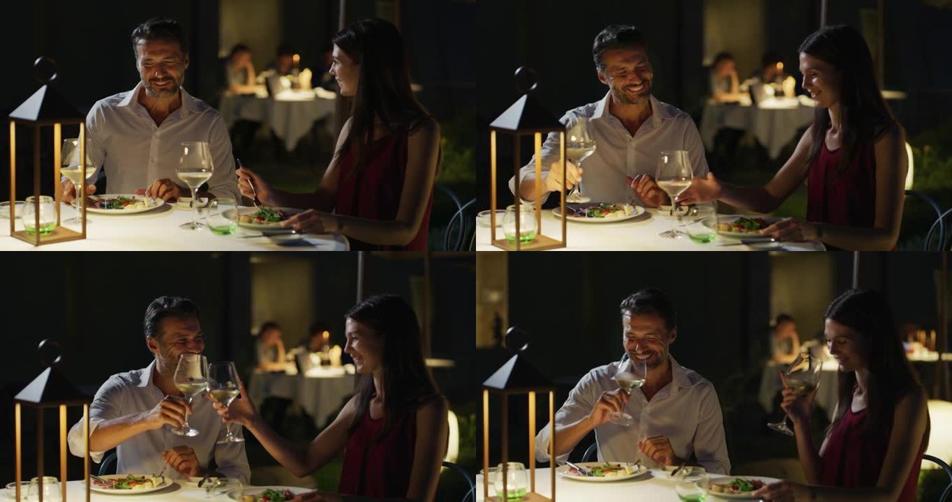 穿着优雅的幸福夫妇一起享受浪漫的晚餐，并在豪华餐厅的餐桌上用白酒杯欢呼，庆祝他们的周年纪念日和永恒的