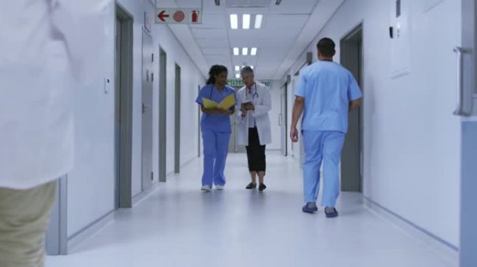 两名不同的女医生走在繁忙的医院走廊上，看着平板电脑和文件，说话