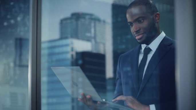 成功的黑人商人的肖像穿着西装站着，用笔记本电脑看着窗外。从事计算机工作的股票市场投资经理电子商务初创