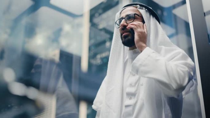 成功的穆斯林商人在传统的白色坎杜拉骑着玻璃电梯到现代商务中心的办公室。男子在电梯里打电话。沙特、阿联