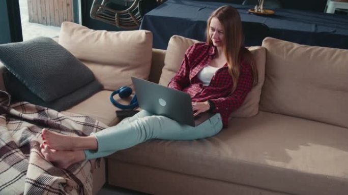 远程工作。美丽严肃专注的年轻白人女商人在家里舒适的沙发上在笔记本电脑上打字信息。