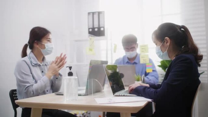 亚洲女商人戴着口罩，用酒精凝胶清洁手部消毒，然后开始在办公室用笔记本电脑工作