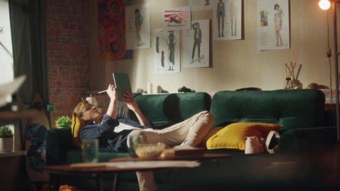 白人男性设计师在数字平板电脑上绘画，同时以轻松的姿势躺在沙发上。阁楼现代房间。聪明的人与新一代概念