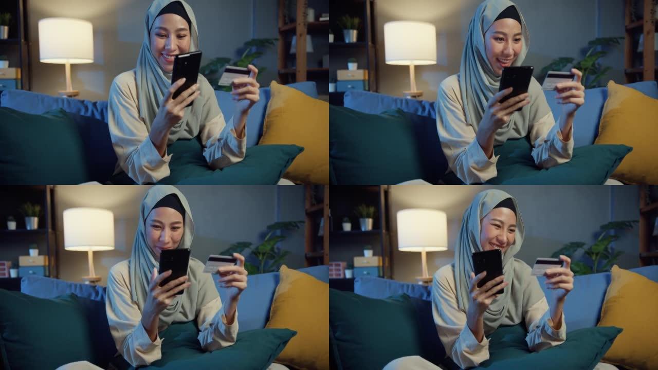 迷人开朗的年轻亚洲伊斯兰穆斯林美女，戴着头巾，随意微笑，坐在沙发上使用电话，晚上在舒适的客厅用信用卡