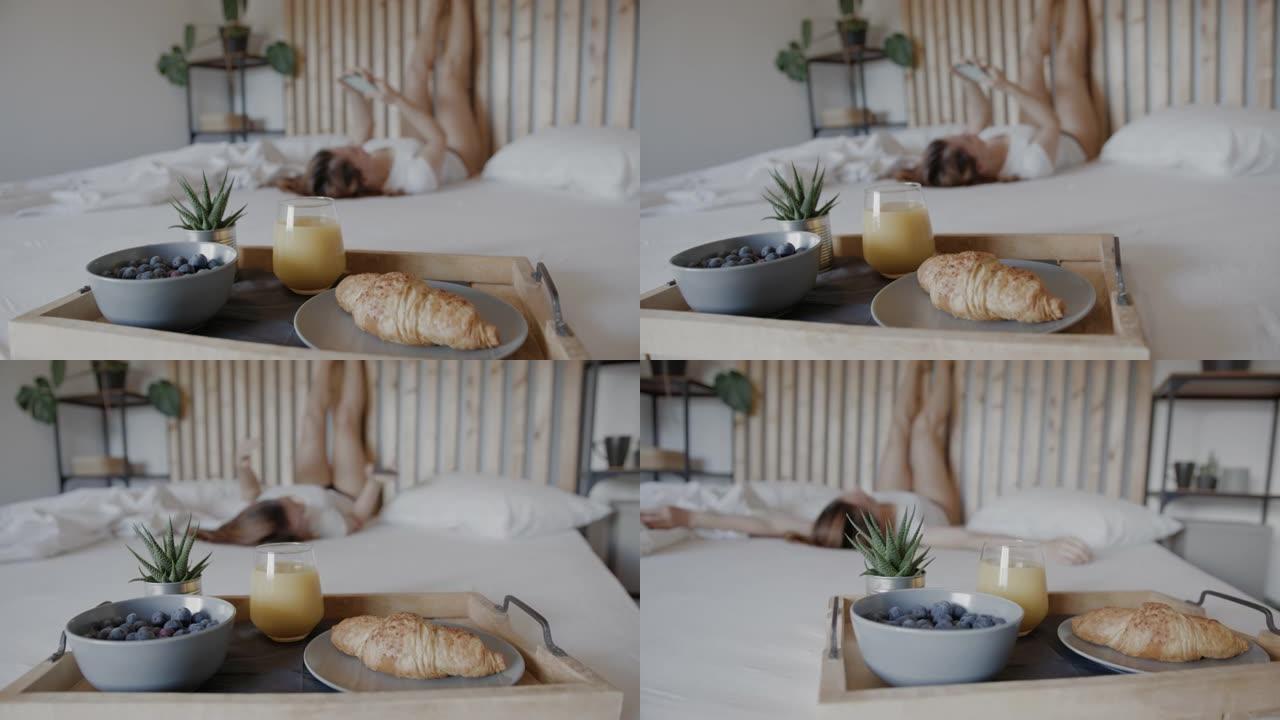 早餐托盘在床上，女人在背景中使用手机，同时将腿靠在墙上