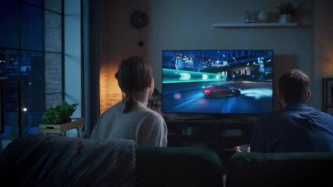 年轻夫妇在家里度过时光，坐在阁楼公寓的沙发上，在游戏机上玩街机汽车视频游戏。男朋友在玩漂移模拟器，而