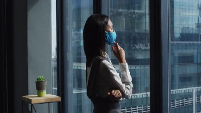 体贴的亚洲妇女戴着口罩在现代办公室看着窗外
