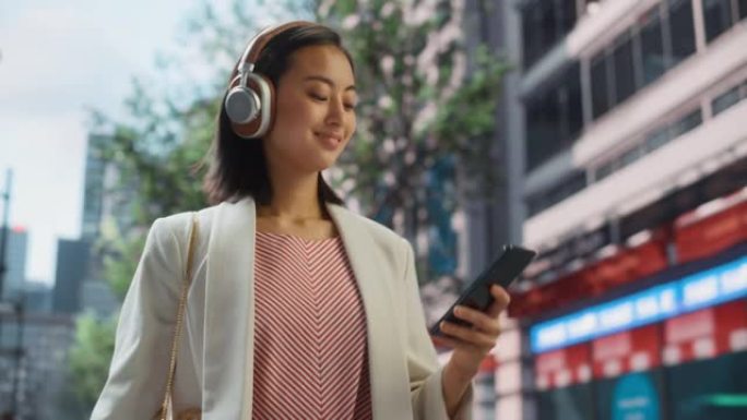 一位美丽的日本女性的肖像随便走在街上，用智能手机的耳机听音乐。她在大城市中使用电话，向人们发送在线消