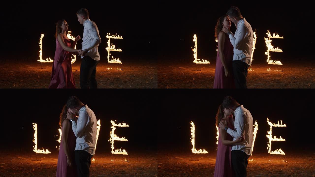 一对年轻深情的夫妇在夜晚亲吻的肖像，背景中的爱这个词闪耀着火焰。男人和女人庆祝自己的周年纪念日，大字