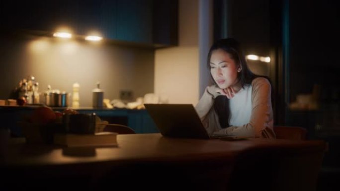 美丽地道的亚洲女人坐在舒适的厨房里的桌子旁，晚上在家使用笔记本电脑。女性在互联网和社交媒体上微笑聊天