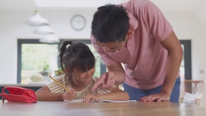亚洲父亲帮助在家上学的女儿在厨房的餐桌上写书