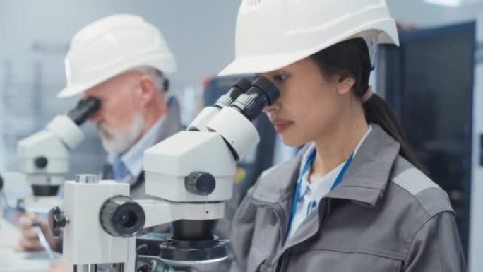 年轻的亚洲工业科学家和年长的工程师在工厂的办公桌上工作，使用显微镜检查制造生产零件的质量。