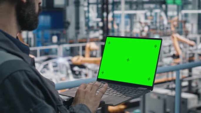 汽车工厂工程师在工作制服中使用带有绿屏模型显示的笔记本电脑。在专门用于车辆生产的汽车工业制造工厂使用