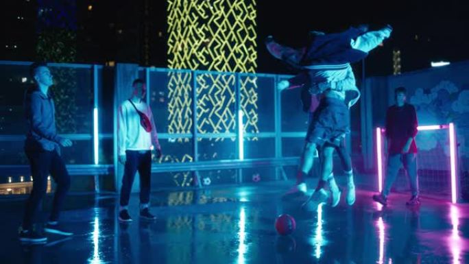 雨后快乐的多元朋友在屋顶城市的户外玩足球，霓虹灯。两名玩家庆祝一场成功的比赛，撞胸部并互相拥抱