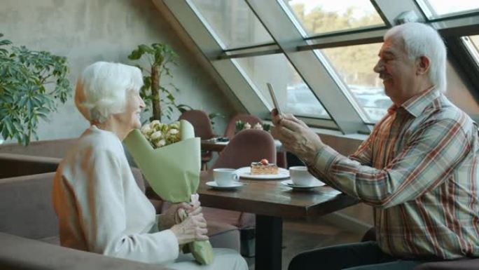 优雅的高级女子与丈夫在咖啡馆共进晚餐时，鲜花为智能手机相机摆姿势