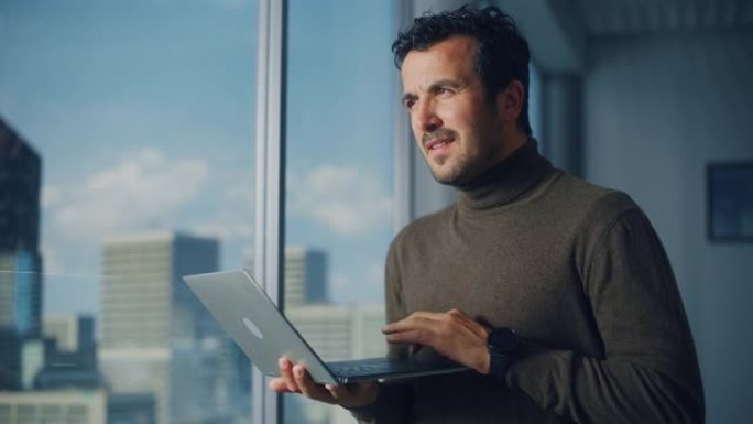 专业的中东商人站在大城市窗户附近的办公室时使用笔记本电脑。成功的软件工程师为他的电子商务创业公司进行