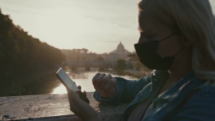 日落时，戴着面具的SLO MO女人在罗马的一座桥上使用智能手机