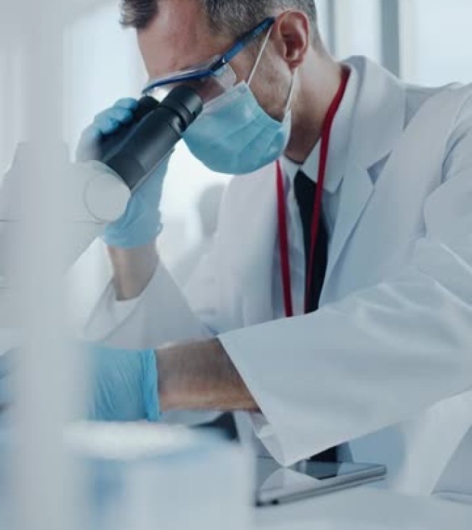 垂直视频。医学发展实验室: 科学家戴着口罩在显微镜下观察，将数据输入数字平板电脑。医学，先进实验室中