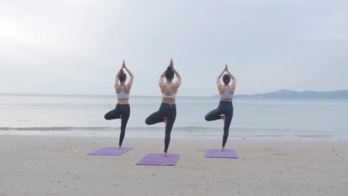 后视图，三人一组体育课在日出的早晨在海滩上一起练习瑜伽课。积极的生活方式理念。