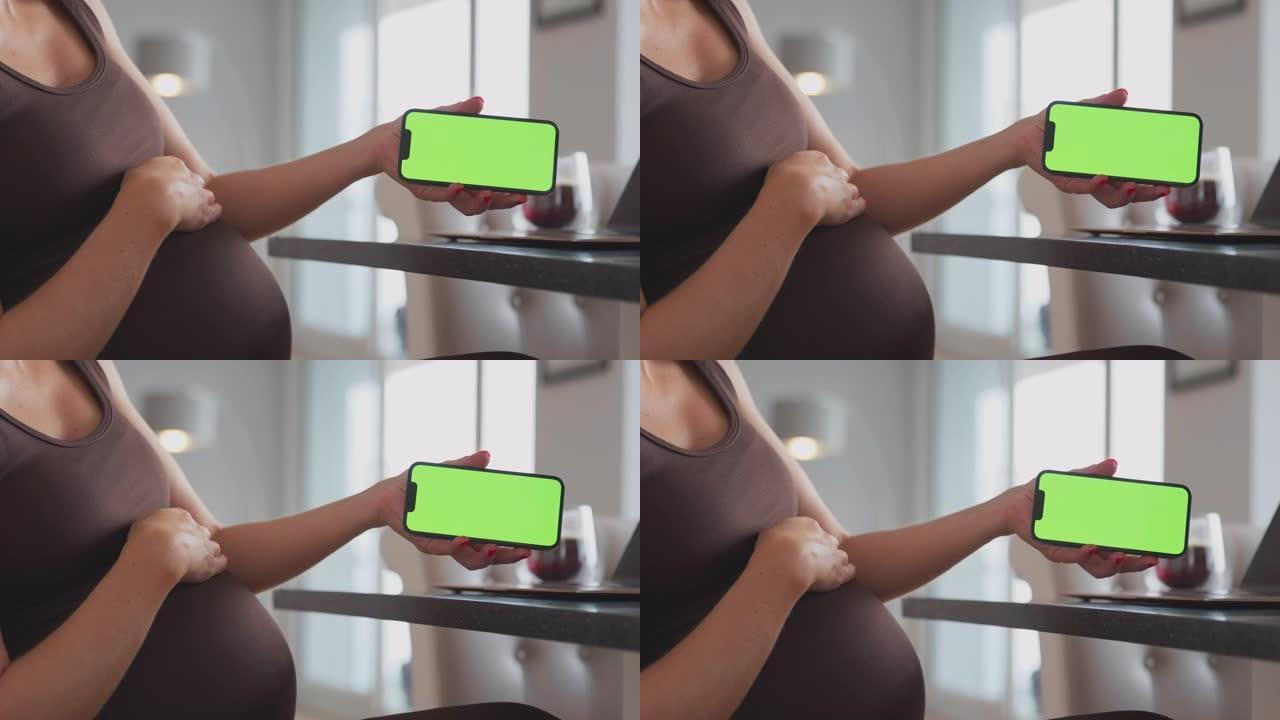 孕妇在家拿着绿屏手机的笔记本电脑上工作的特写