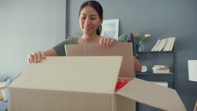 快乐美丽的亚洲女士从客厅的在线市场上拆箱纸板递送包。