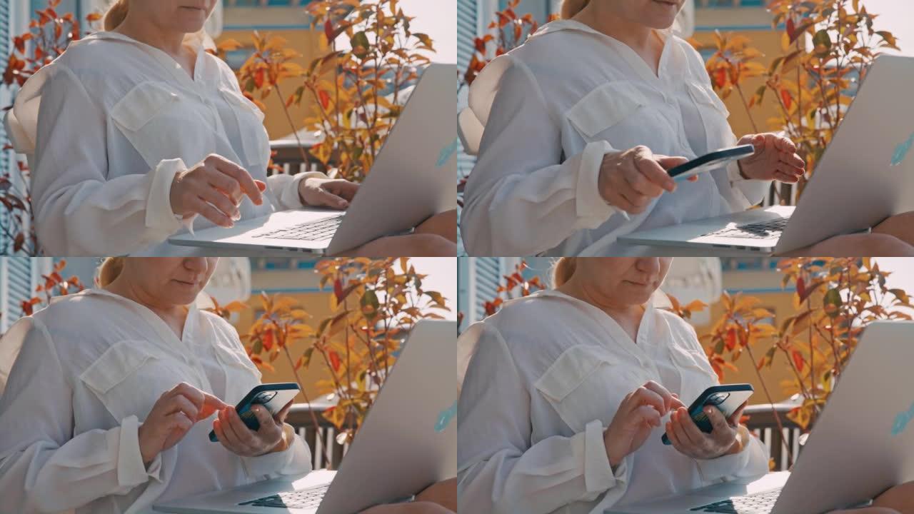 金发碧眼的女人坐在酒店房间的阳台上，在笔记本电脑上工作，被智能手机上的警报分散了注意力