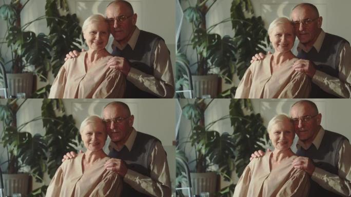 幸福可爱的浪漫70年代高级夫妻肖像，退休夫妇拥抱，微笑着看着相机在家