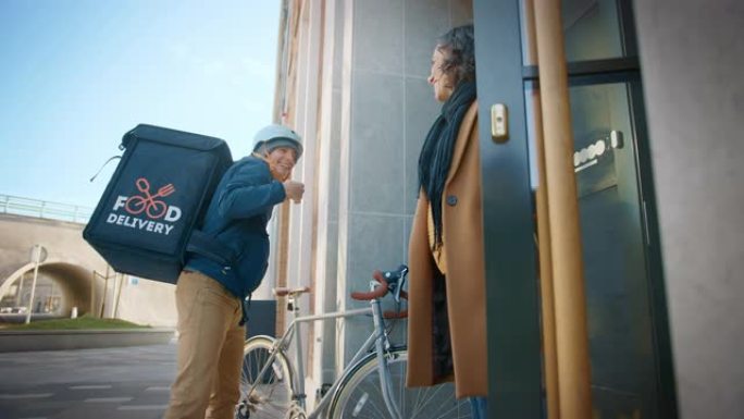 快乐的送餐男子在自行车上穿着保暖背包，向一位美丽的女性顾客提供披萨订单。Courier在办公楼里给一