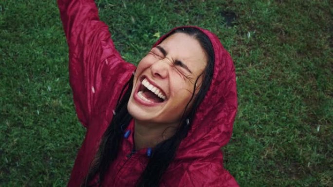 高角度拍摄:穿着红色雨衣的快乐女人的肖像，雨水落在她的脸上微笑和大笑。被绿草包围的成年女性与她内心的