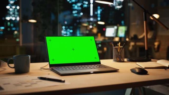 带有绿屏Chromakey模拟显示器的笔记本电脑晚上站在办公室的桌子上。现代开放空间创意机构，经理在