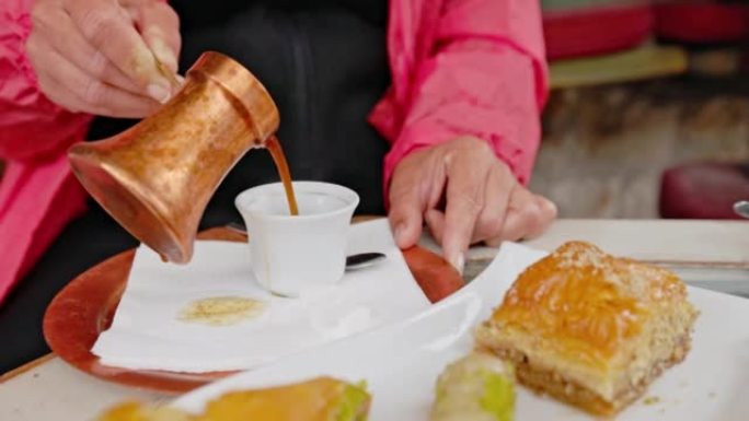 特写女人，穿着红色外套，在早餐时从青铜色的锅中装满咖啡，蛋糕站在前面的盘子上