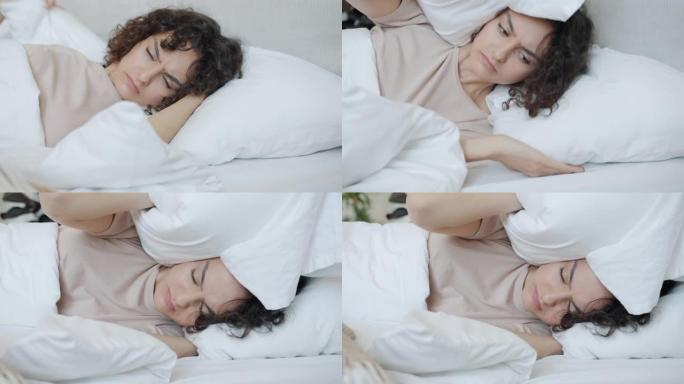 不快乐的年轻女子醒来，用枕头遮住耳朵，讨厌噪音