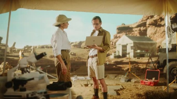 考古挖掘现场: 两位伟大的考古学家站在发掘现场的帐篷工作中，使用笔记本电脑检查新发现的古代文明建筑遗