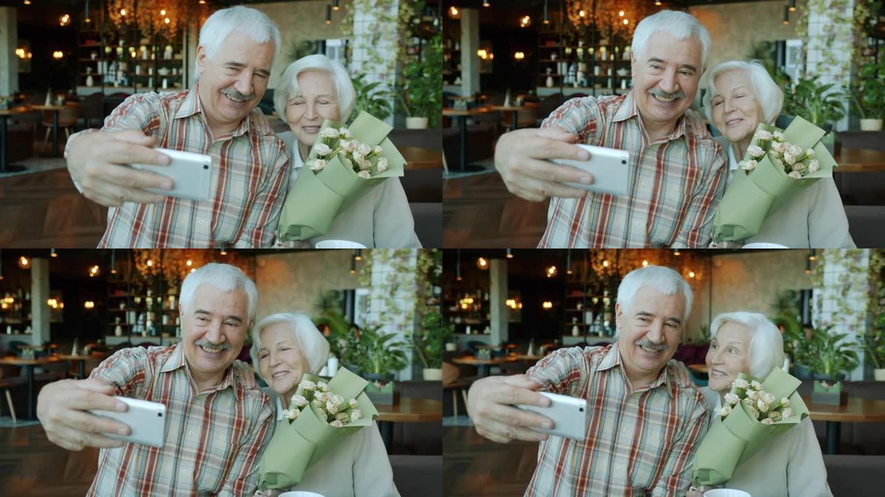 美丽的老年夫妇在咖啡馆用手持鲜花的智能手机自拍