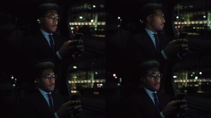 时髦的戴眼镜的黑人在晚上坐出租车的后座上下班。穿着西装的英俊男性在城市街道上使用智能手机，在汽车上看