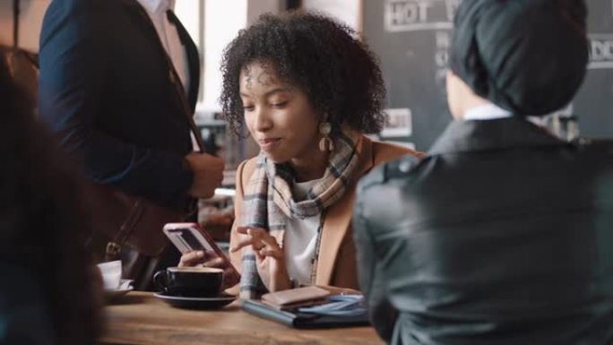 美丽的非洲裔美国妇女在咖啡馆使用智能手机发短信在社交媒体上分享信息，享受移动技术在繁忙的餐厅与朋友聊